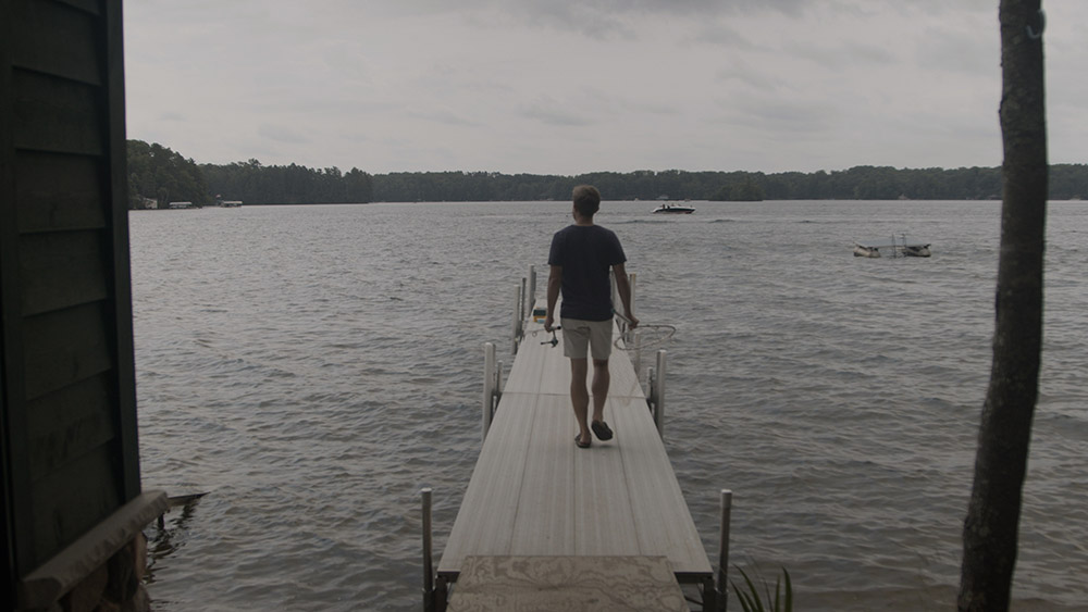 Young man in blue shirt and khaki shorts, walking down a dock with a fishing pole in his left hand