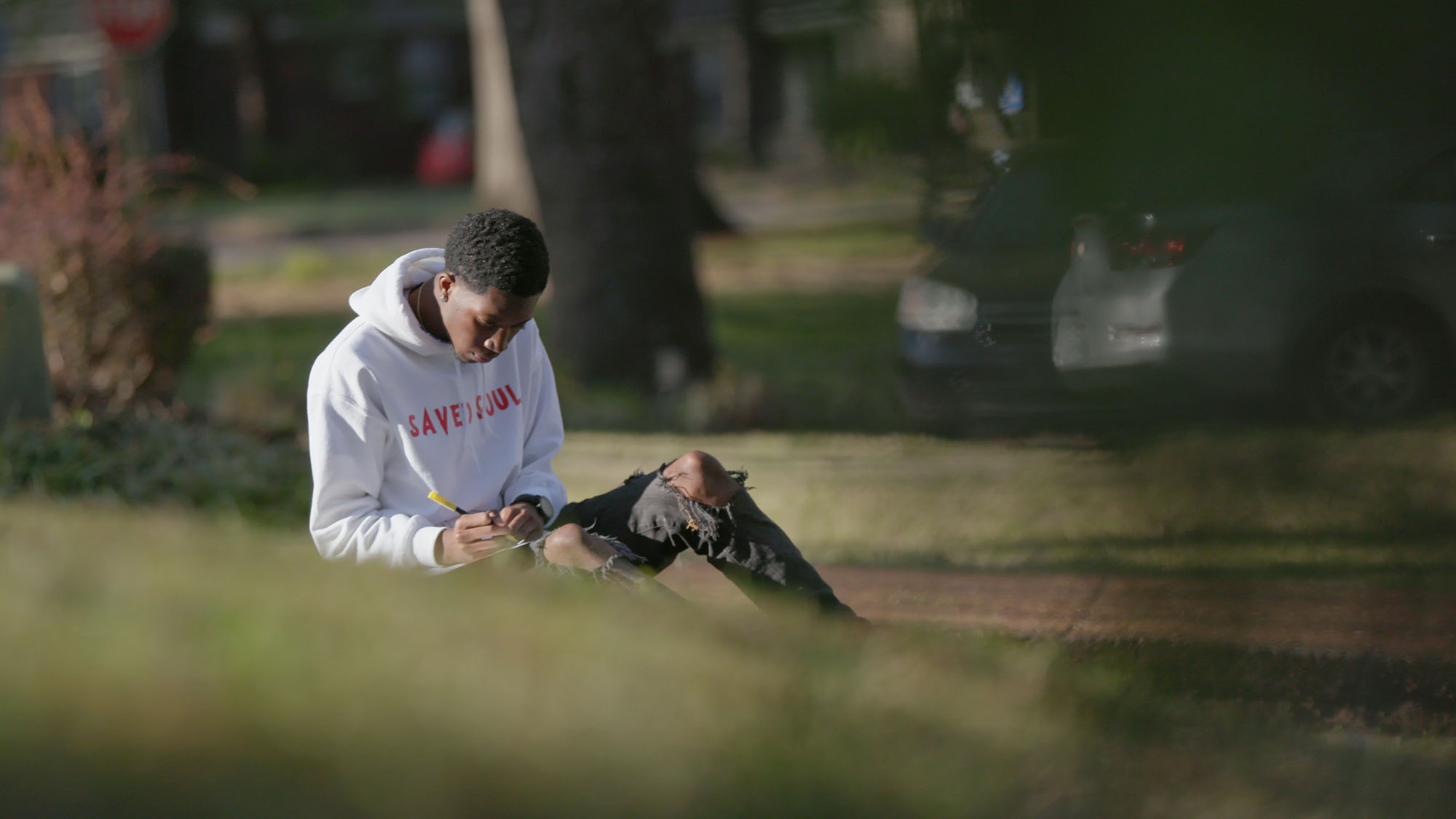 Young man in a white hoodie, sitting in park, drawing in a notebook.