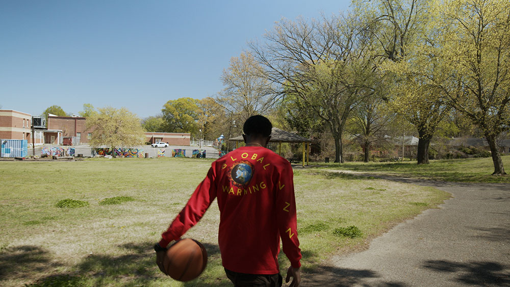 Kenneth Bell - wearing a red Global Warming shirt, walking away from the camera dribbling a basketball