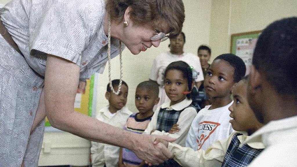 Janet Reno as attorney general, in a classroom shaking hands with a young child in a school uniform